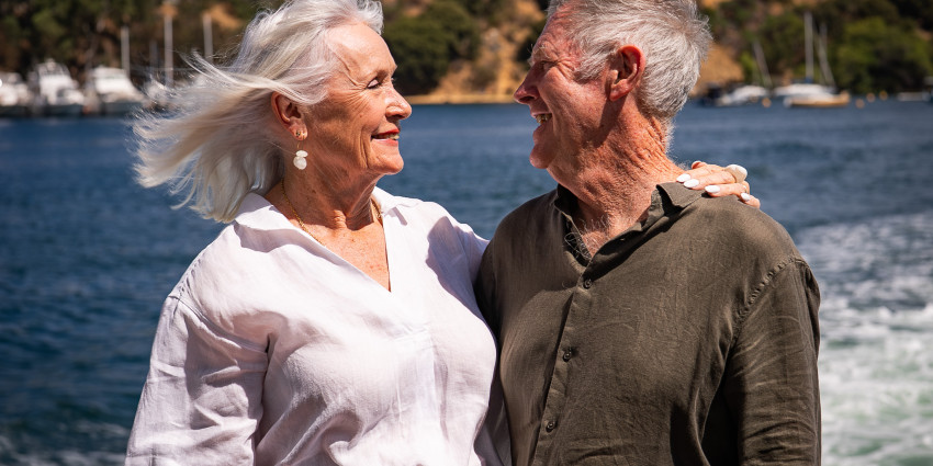 An older man and woman smiling, hugging, with the ocean in the background