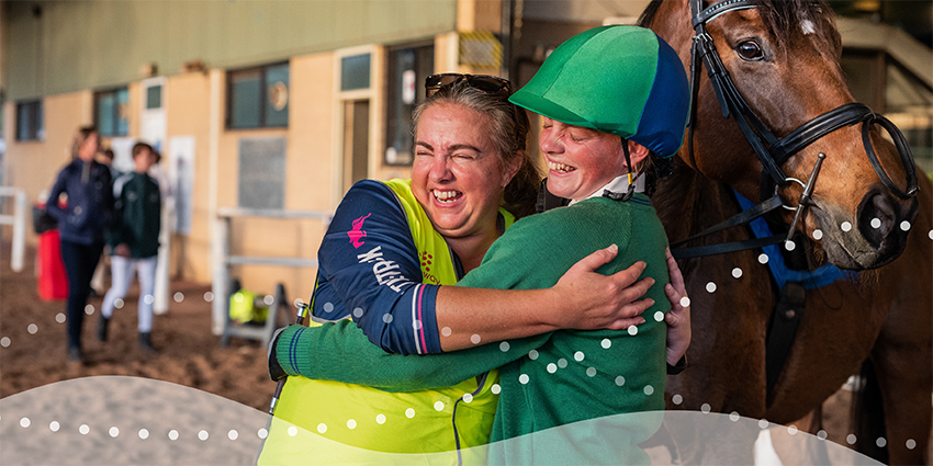 two adult volunteers smiling and hugging, with a horse in the background