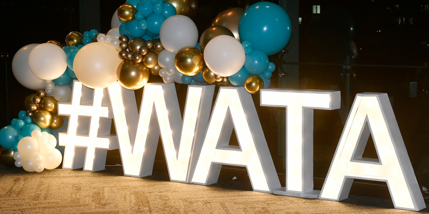 Illuminated “#WATA” sign with teal, white, and gold balloon decorations at an indoor event.
