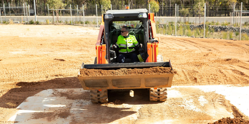 Person operating a skid steer loader carrying soil across a dirt training site.