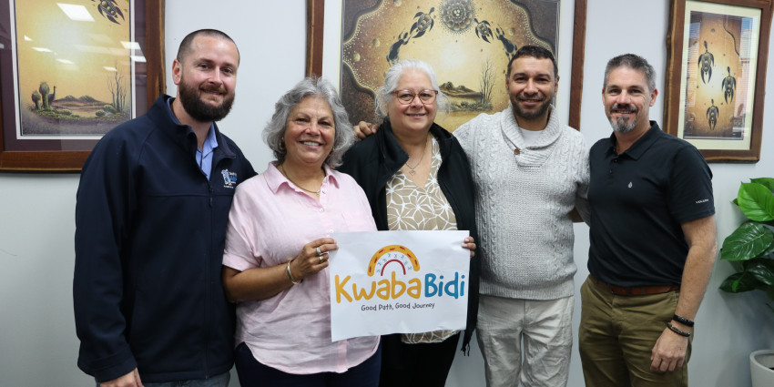 Five people stand together in an office beneath three framed artworks. Two people in the centre hold a sign reading “Kwaba Bidi – Good Path, Good Journey.” The group is posed for a photo against a white wall with Indigenous-style artwork.