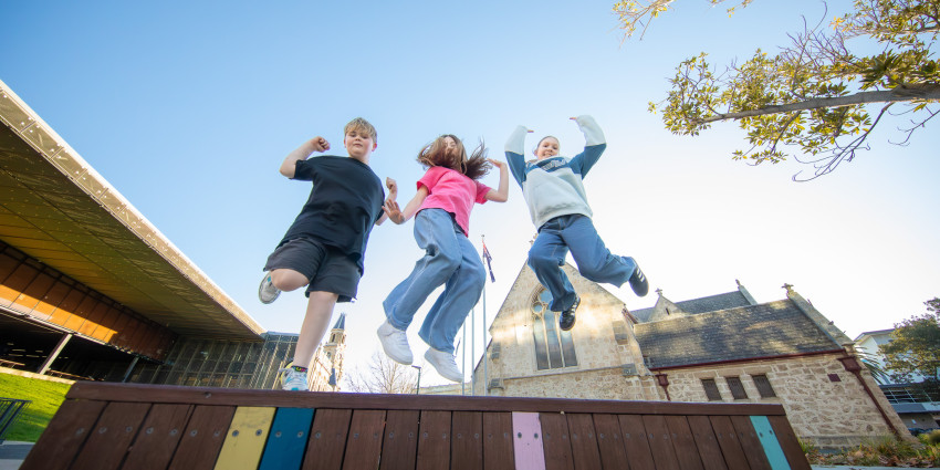 Three children jump off a colourful wooden bench in an outdoor plaza, with a historic stone building, modern architecture, and clear blue sky in the background.