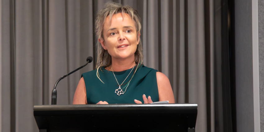 Caroline Speirs speaking at a lectern with a microphone in front of grey curtains at the Child Protection Excellence Awards.