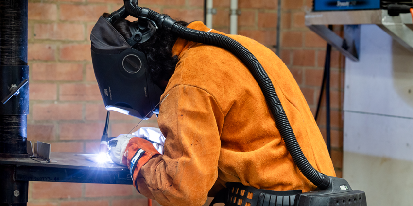 Student welding metal in a workshop while wearing protective safety gear.