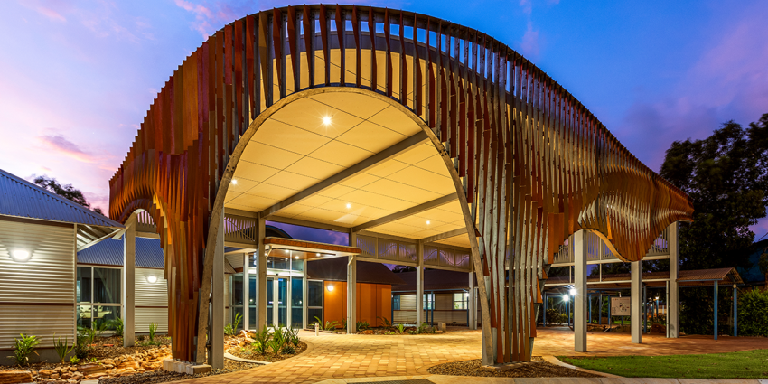 Entrance to a North Regional TAFE campus featuring a distinctive curved steel canopy, illuminated at dusk with landscaped gardens and paved walkways.