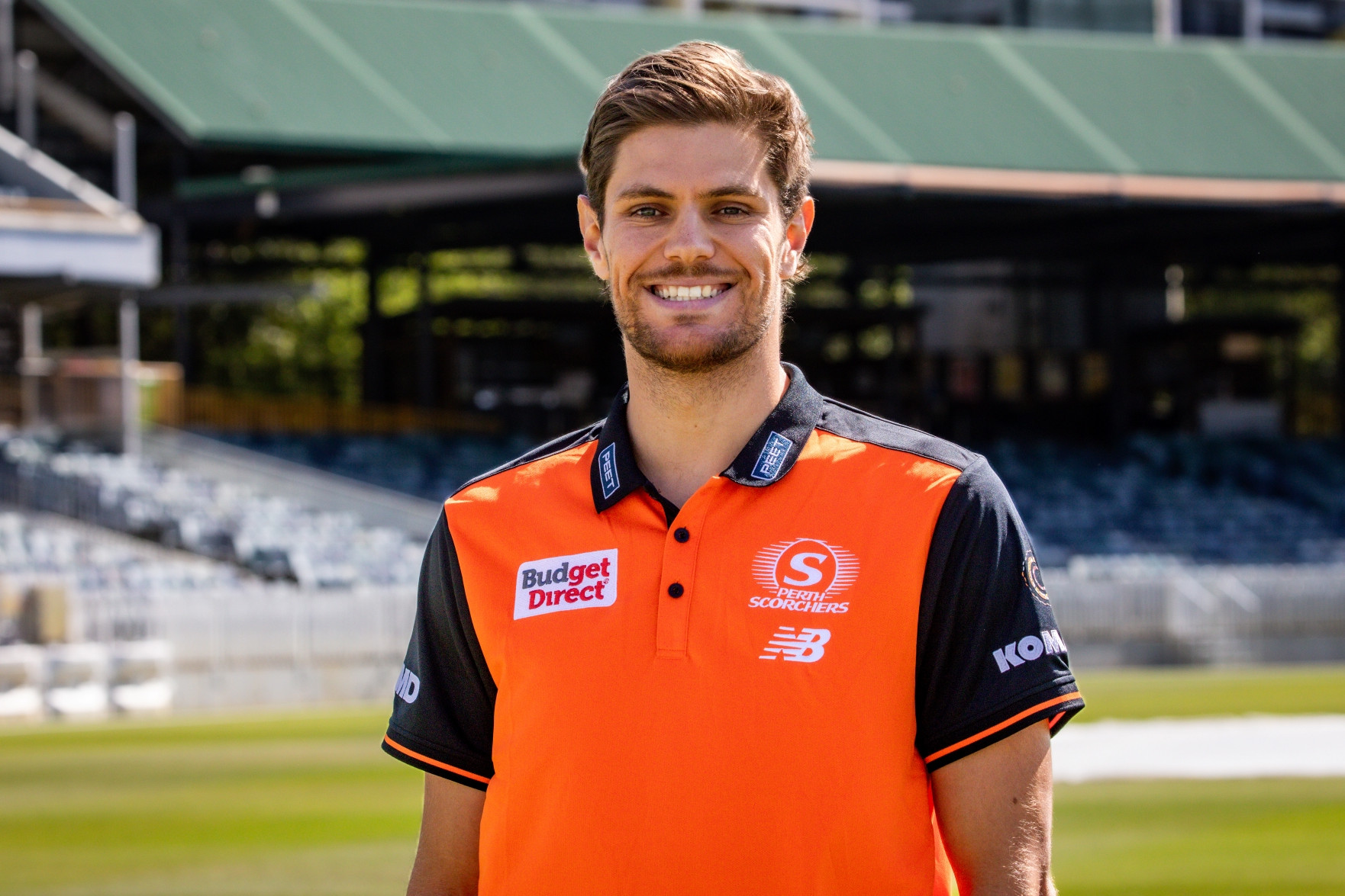 a sportsman in an orange sports top standing in a cricket oval