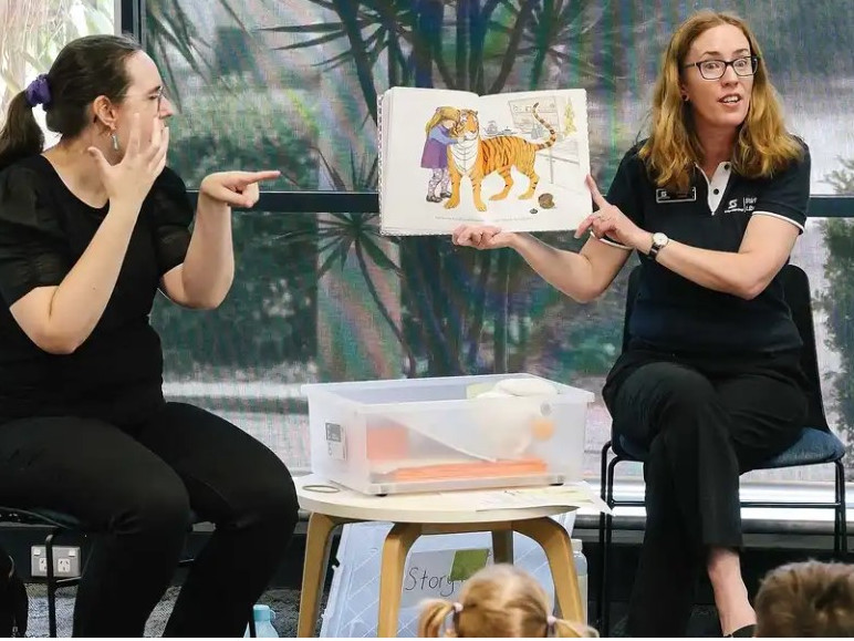 Two young women presenting a children's story book to children, including in Auslan sign language.